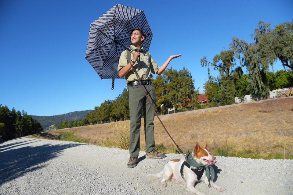 Flood Ready Freddy holding an open umbrella over his shoulder and a dog leash in his right hand. Medium white and brown dog laying on gravel road