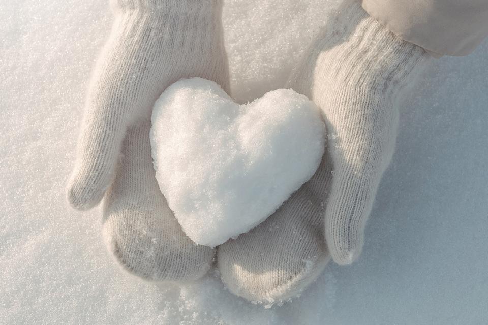 A child wearing white mittens delicately holding a heart-shaped snowball against a snowy background